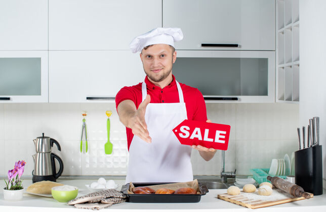 观点顶视图年轻微笑的男厨师手持销售标志 在白色厨房欢迎某人烹饪成人男性厨师