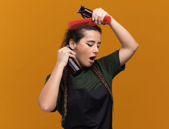 感情印象深刻的年轻女理发师在统一举行理发工具和梳头孤立在橙色墙上立场表情姿势