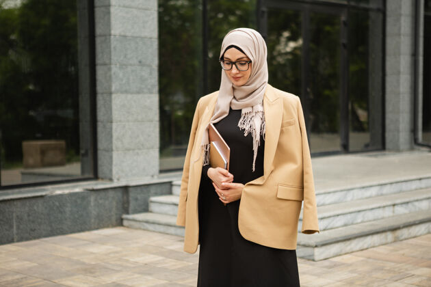 自营职业现代时尚的妇女戴着头巾 穿着商务风格的夹克和黑色的长袍 带着笔记本电脑走在城市街道上就业时尚围巾