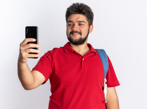 年轻年轻的留着胡子的学生 穿着红色马球衫 背着背包 手里拿着智能手机 站在白色的墙上兴高采烈地笑着看着它移动背包欢呼