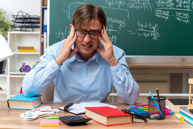 黑板戴眼镜的年轻男老师坐在教室的黑板前 手里拿着书和笔记 紧张而紧张穿着紧张教室
