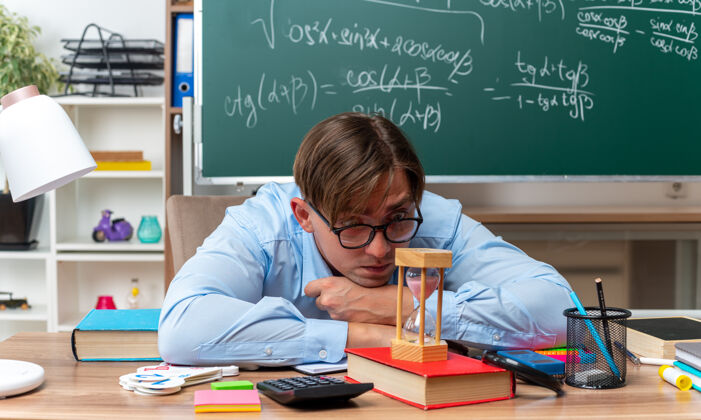 年轻年轻的男老师戴着眼镜看着沙漏疲惫而无聊地坐在课桌旁 教室里的黑板前放着书和笔记无聊教室黑板