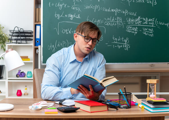 笔记年轻的男老师戴着眼镜看书备课看起来很自信地坐在课桌旁 教室里的黑板前放着书和笔记课桌黑板年轻