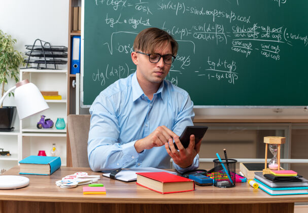 教室年轻的男老师戴着眼镜用智能手机打字 看起来很自信地坐在课桌旁 教室的黑板前放着书和笔记笔记留言年轻