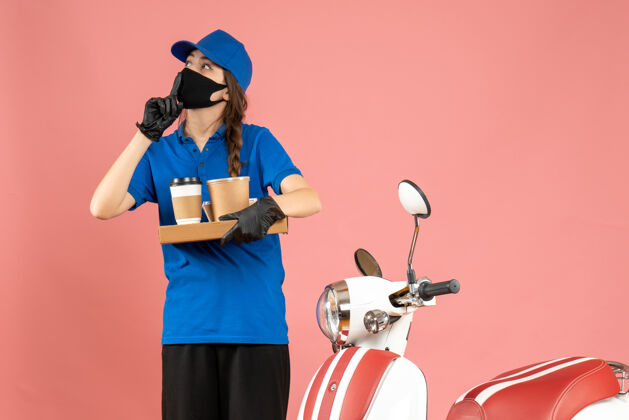 焦点俯视图：戴着医用面罩手套的快递员女孩站在摩托车旁 手里拿着粉色背景上的咖啡小蛋糕男医药蛋糕
