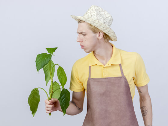 帽子年轻的园丁穿着连体衣戴着帽子拿着植物看着它困惑连身衣花园困惑