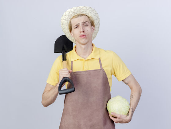 连身衣年轻的园丁穿着连体衣 戴着帽子 手里拿着铲子和卷心菜 看上去很自信穿花园卷心菜