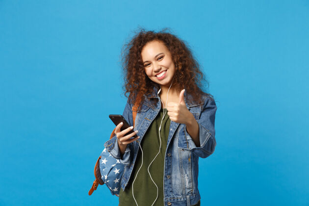 站立年轻的非洲裔美国少女 穿着牛仔服 背着耳机 隔离在蓝色的墙上设备人电话