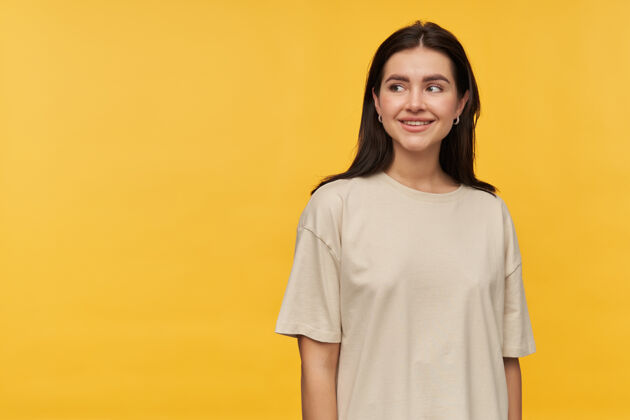 微笑快乐美丽的黑发年轻女子穿着白色t恤站在一旁看着黄色墙壁上的空地人物可爱姿势