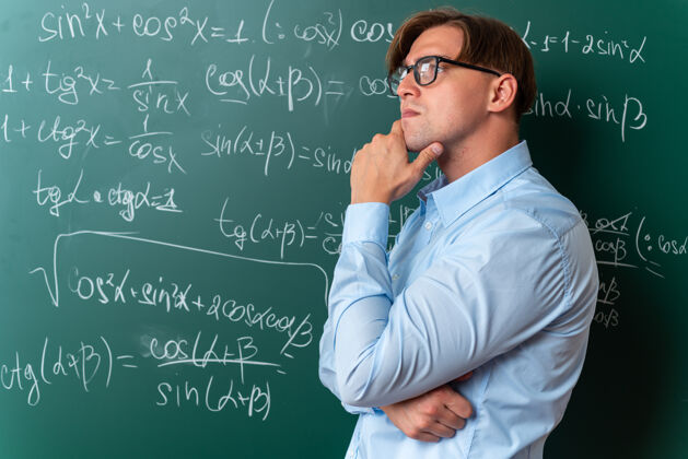 沉思年轻的男老师戴着眼镜 手放在下巴边 沉思着 站在黑板边思考着数学公式 在教室里黑板下巴课堂