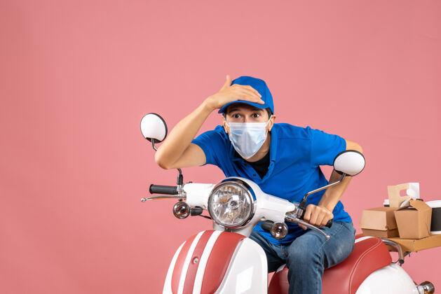 滑板车前视图集中快递员戴着医疗面具戴着帽子坐在粉彩桃色背景的踏板车上面具医学摩托车