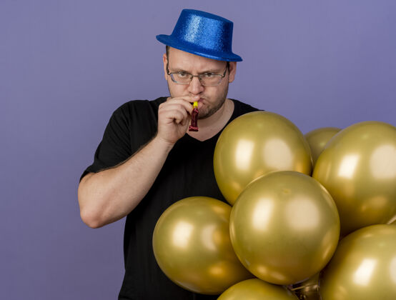 斯拉夫严肃的成年斯拉夫人戴着眼镜 戴着蓝色的派对帽 站着氦气球吹派对哨子复制派对眼镜