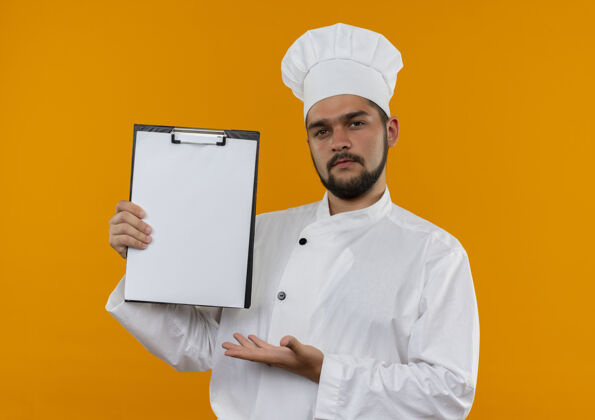 厨师自信的年轻男厨师身着厨师制服 用手指着橘色墙上的剪贴板显示烹饪制服