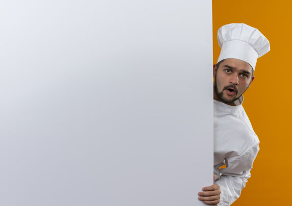 墙令人印象深刻的年轻男厨师穿着厨师制服从后面看 把手放在白色的墙上隔离在橙色的墙上与复制空间厨师烹饪制服