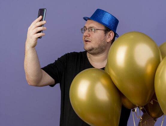 成人给人印象深刻的成年斯拉夫男子戴着眼镜 戴着蓝色派对帽 站着氦气球看手机空间斯拉夫氦