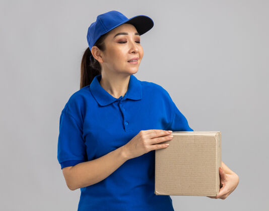 表情身穿蓝色制服 戴着帽子的年轻送货员站在白色的墙上 带着自信的表情看着旁边的纸板箱女人制服年轻