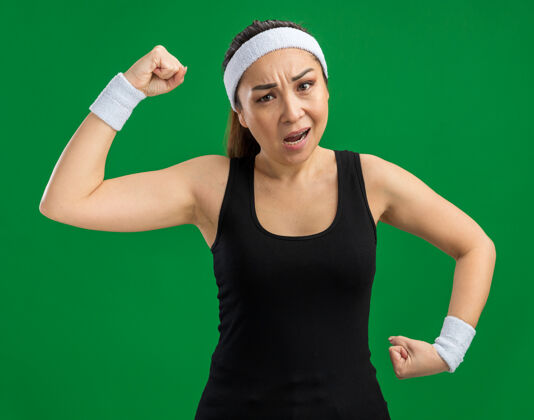 力量戴着头带和臂章的年轻健身女士绷紧了 自信地举起拳头 站在绿色的墙上 展现出力量和力量年轻站立紧张
