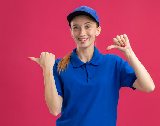 信心身穿蓝色制服 头戴鸭舌帽的年轻送货员微笑着自信地用拇指指着站在粉色墙上的一侧女孩立场拇指