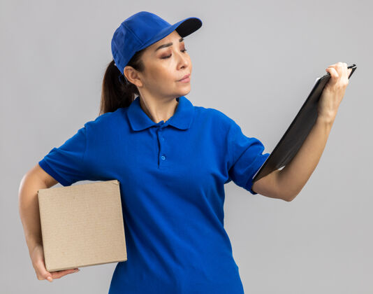 严重身穿蓝色制服 头戴帽子的年轻送货妇女拿着纸板箱和剪贴板 严肃地站在白色的墙上看着它女人制服举行