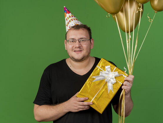 复制带着眼镜 戴着生日帽 面带微笑的成年斯拉夫男子手持氦气球和礼盒盒子空间成人