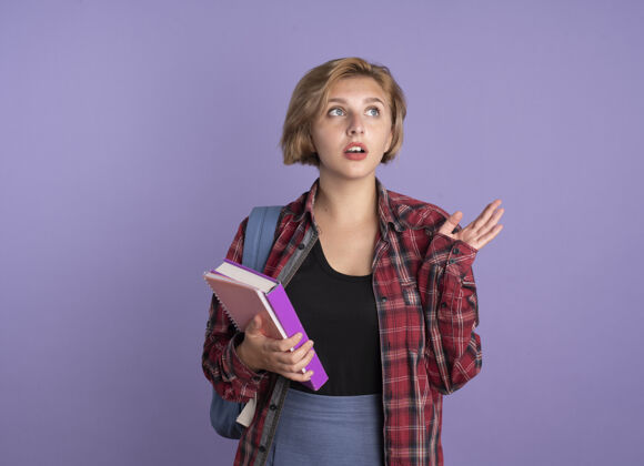笔记本焦急的年轻斯拉夫学生女孩穿着背包站着 举起手拿着书和笔记本看着身边提高斯拉夫焦虑