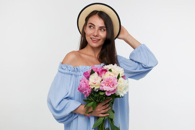 成人有魅力的女孩的肖像 长黑发戴着帽子和蓝色漂亮的裙子拿着一束花和触摸一顶帽子花束完美美丽