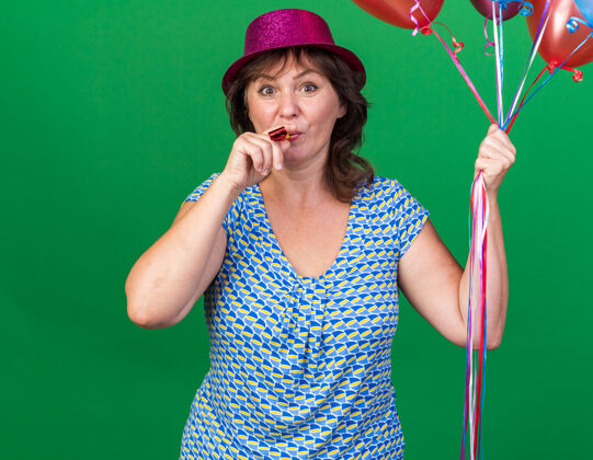 五颜六色快乐的中年妇女戴着派对帽手持五颜六色的气球吹着口哨快乐的生日派对站在绿色的墙上年龄立场举行