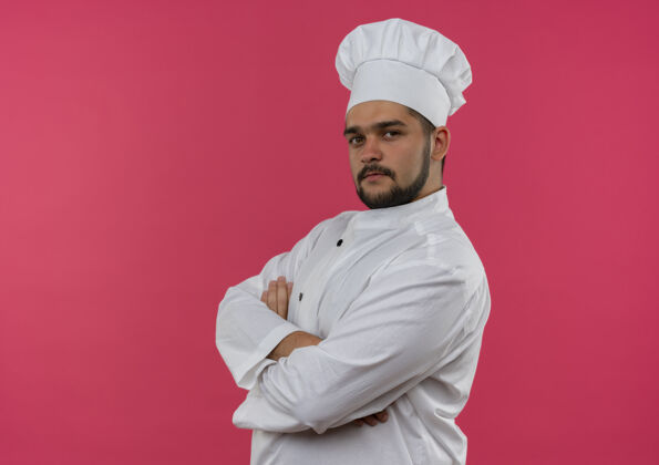 烹饪自信的年轻男性厨师身着厨师制服站在侧面视图中 封闭的姿势隔离在粉色墙壁上 留有复制空间视野制服站立