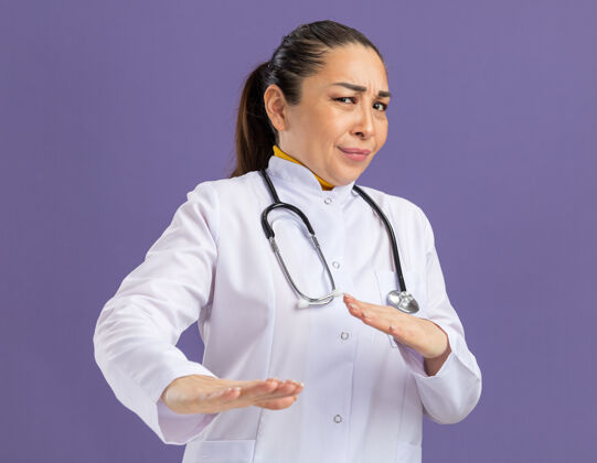 站年轻的女医生穿着白药衣 脖子上戴着听诊器 表情厌恶 双手站在紫色的墙上做着防御的手势防御表情外套