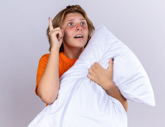 流感不健康的年轻女子穿着橙色t恤 抱着枕头 感觉不舒服 患流感 抬头惊讶地看着 食指有了新的想法站在白色的墙上年轻手指感情