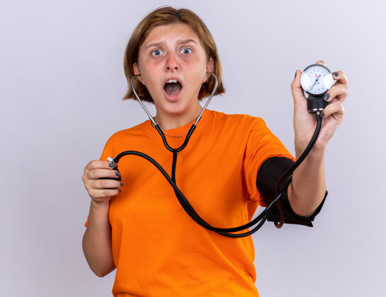 站立不健康的年轻女子穿着橙色t恤 感觉不舒服 站在白色的墙上用血压计测量血压 看起来很担心 很害怕不健康压力感觉