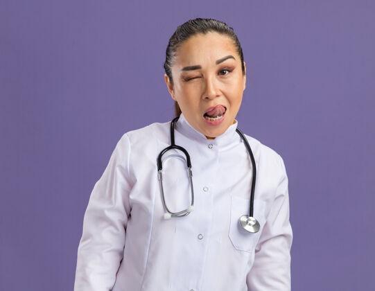 棒快乐的年轻女医生 穿着白药衣 脖子上戴着听诊器 站在紫色的墙上眨眼 伸出舌头女人眨眼外套