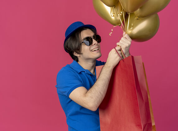 纸一个戴着太阳眼镜 戴着蓝色派对帽的快乐英俊的白种人拿着氦气球和纸购物袋帅哥穿眼镜