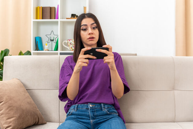 使用穿着休闲服的小女孩坐在客厅的沙发上玩着智能手机游戏 看起来很自信坐着女人休闲