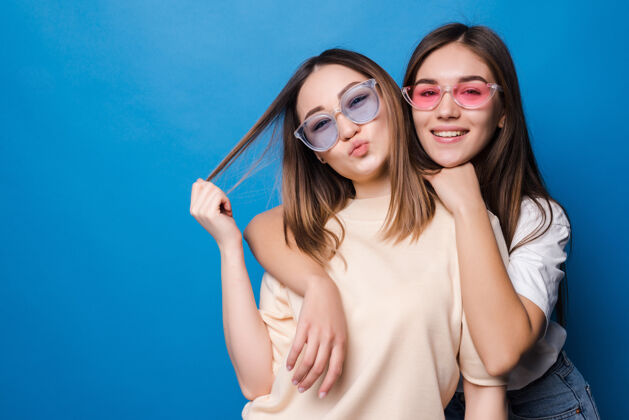 时尚永远的朋友两个戴着太阳镜的可爱女孩在蓝色的墙上微笑着摆姿势乐趣快乐女人
