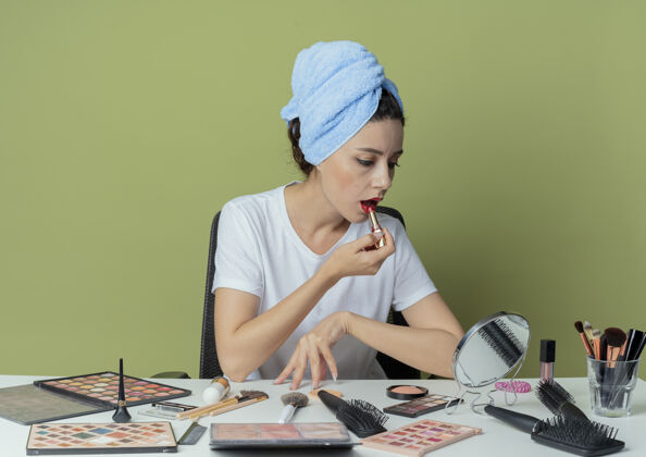 女孩年轻漂亮的女孩坐在化妆台上用化妆工具和毛巾在头上看着镜子 戴上红唇膏 在橄榄绿的空间里触摸桌子工具化妆品触摸