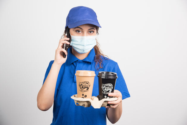 穿着蓝色制服 戴着医用口罩的女人在打电话 手里拿着两杯白咖啡面罩帽子黑发