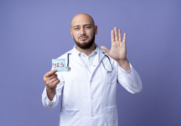 医学年轻的秃头男医生 穿着医用长袍 手持听诊器 手持纸印 做着停止的手势男性显示停止
