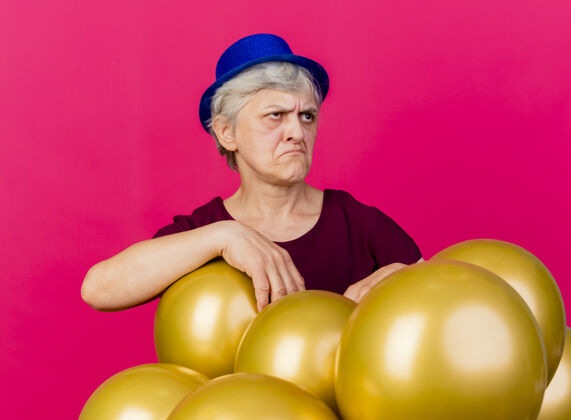 女人心烦意乱的老妇人戴着派对帽站在氦气球旁边看着粉红色的站穿聚会