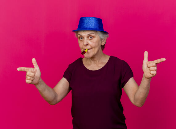 女人惊讶的老妇人戴着聚会的帽子指着两边吹着粉红色的哨子点党帽子
