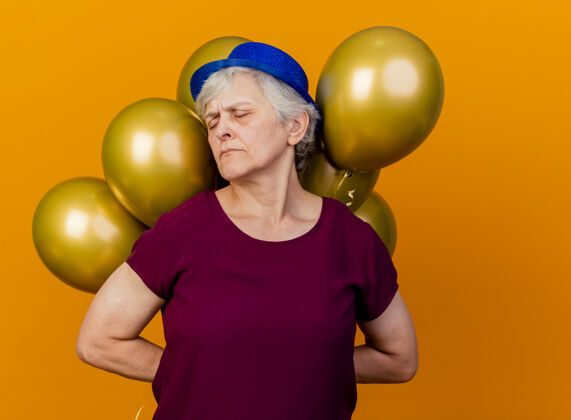 气球一位戴着派对帽的老妇人 紧闭着眼睛 身后拿着氦气球 注视着橙色女人帽子派对