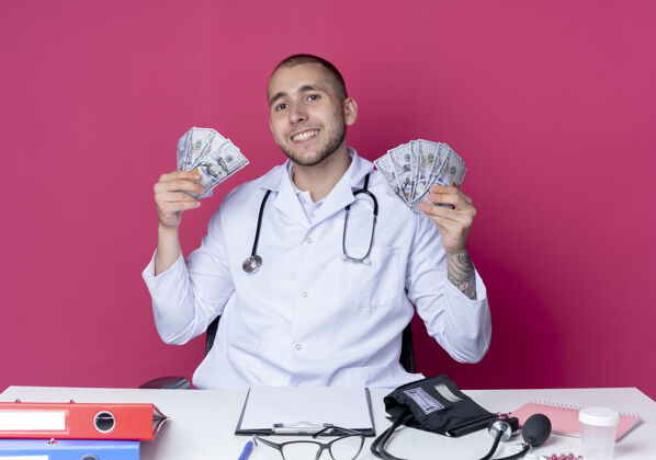 桌子面带微笑的年轻男医生穿着医用长袍和听诊器坐在办公桌旁 手里拿着工作工具 手里拿着隔离在粉红色墙上的钱表情脸姿势