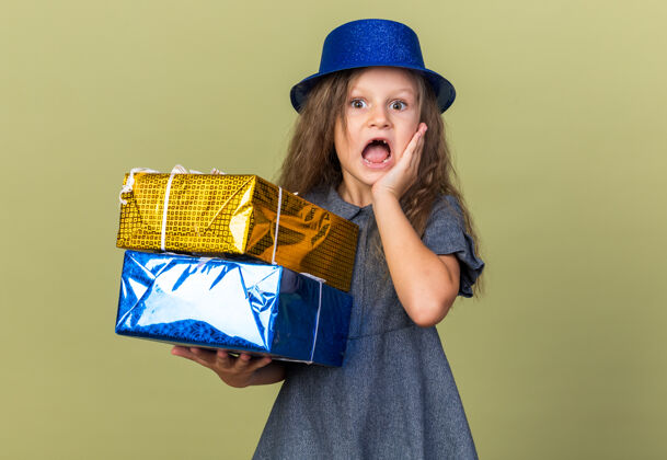 聚会焦急的金发小女孩 戴着蓝色派对帽 手放在脸上 手拿着礼盒 孤立地放在橄榄绿的墙上 还有复印空间女孩橄榄生日