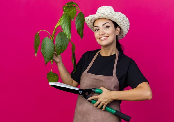 表情年轻漂亮的女园丁穿着围裙 戴着帽子 拿着植物和修剪篱笆的剪子 站在粉红色的墙上 面带微笑地看着前面篱笆人帽子