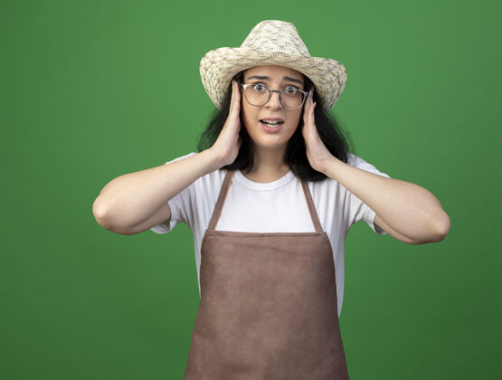 姿势焦急的年轻黑发女园丁戴着眼镜和制服 戴着园艺帽 手放在隔离在绿色墙上的脸上手焦虑年轻人