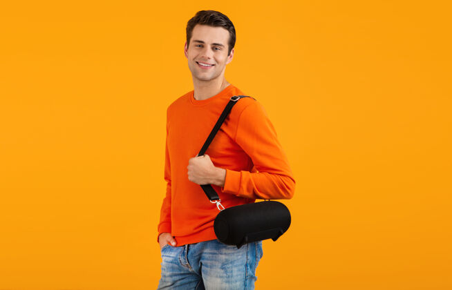 聚会穿着橙色毛衣的时髦微笑的年轻人拿着无线扬声器快乐地听音乐享受橙色的乐趣娱乐肖像家伙