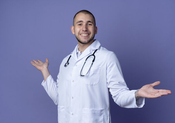手面带微笑的年轻男医生 身穿医用长袍 脖子上戴着听诊器 双手空空地孤立在紫色的墙上人年轻脖子
