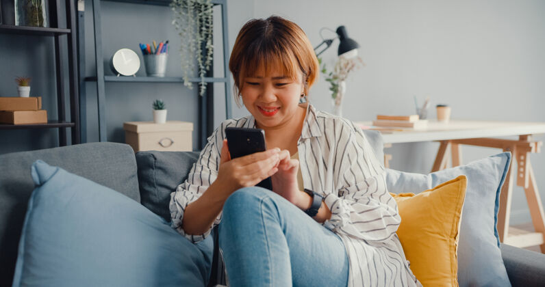 女人年轻的亚洲女士在客厅的沙发上用智能手机发短信或查看社交媒体房子自信护理