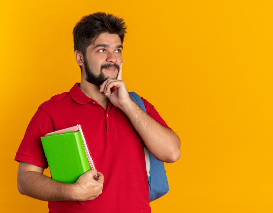 旁白年轻的留着胡子的学生 穿着红色马球衫 背着背包 拿着笔记本 一边沉思 一边思考 一边微笑着站在橙色的背景上小伙子胡子马球