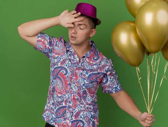 男人疲惫的年轻人闭着眼睛戴着粉红色的帽子拿着气球把手腕放在额头上隔离在绿色气球绿色封闭